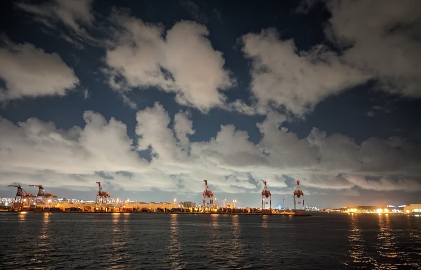Container cranes lit along a bay at night, under a broken cloud sky.
